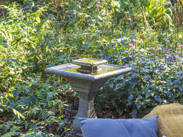 A shapely stone fountain is nestled in abundant native perennials, overlooking a an outdoor sofa.